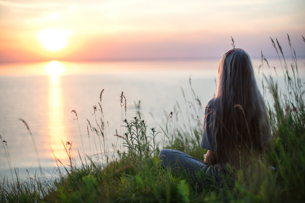 woman sitting on a cliff surrounded by foliage and staring at a large body of water